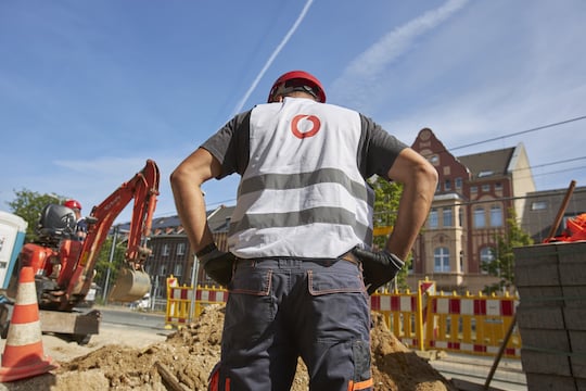 Rckwrtige Ansicht eines Vodafone-Bauarbeiters mit rotem Bauhelm und wei-grauer Warnweste, auf der das rote Vodafone-Logo zu sehen ist; der Bauarbeiter steht vor einer Baustelle, neben der ein Mini-Bagger anrollt