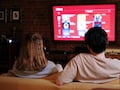 ein Mann und eine Frau sitzen mit dem Rcken zum Betrachter gewand auf einem hellen Sofa und schauen auf einen Fernseher, der ein unscharfes Bild zeigt