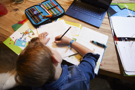 Vogelperspektive auf ein an einem Holztisch sitzendem Kind, dass etwas in ein Schulheft schreibt und dafr einen Stift in der rechten Hand hlt, in der Mitte des Tisches ist ein aufgeklappter schwarzer Laptop zu sehen